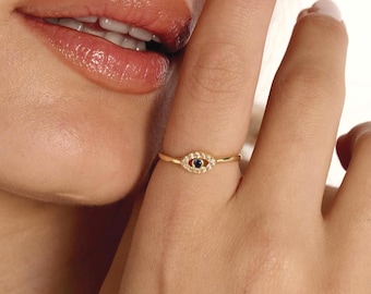 Pode incluir: Um anel dourado com um design de olho. O olho tem um centro preto e é rodeado por pequenas pedras transparentes. O anel é usado num dedo.