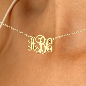 May include: A delicate gold-colored necklace featuring a monogram pendant. The pendant displays three stylized letters in a cursive font. The necklace is worn around the neck, with the chain and pendant visible against the skin.