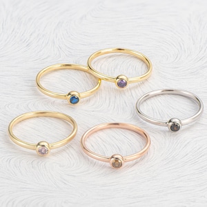 May include: Five simple gold rings with a single gemstone in each. The gemstones are blue, purple, brown, pink, and black.