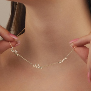 May include: A delicate gold necklace featuring three names: "bella", "chloe", and "steve" in a cursive font. The necklace is held by a person, showcasing the personalized jewelry against a skin tone background.