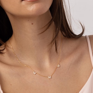 May include: A delicate gold-toned necklace featuring four small butterfly pendants. The butterflies appear to be adorned with small, sparkling accents. The necklace is worn against a light pink top.
