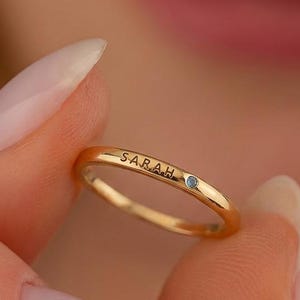 May include: A thin gold band ring featuring the name SARAH engraved in black capital letters on the outer surface, accompanied by a single small, round, light blue gemstone set into the metal next to the name inscription.