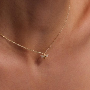 May include: A delicate gold chain necklace features a small, detailed bee pendant adorned with tiny, sparkling clear stones. The bee is intricately designed with visible wings and body segments. The necklace is shown against a brown leather background. The chain is thin and delicate, suitable for everyday wear. The overall aesthetic is minimalist and elegant, perfect for adding a touch of subtle sparkle to any outfit.