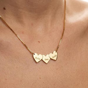 May include: A gold-colored necklace with three heart-shaped pendants. Each heart is engraved with a name: Kylie, Sam, and Emily. The necklace chain has a box link design.