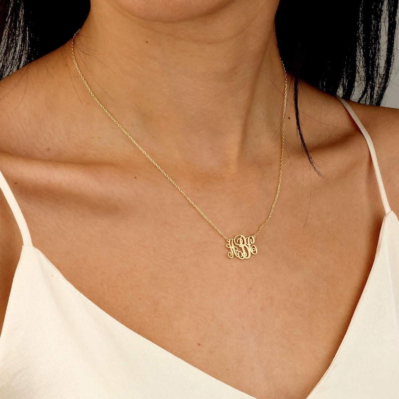 May include: A gold-colored monogram necklace with a delicate chain. The pendant features three intertwined letters in an elegant, cursive font. The necklace is worn around the neck, against a white top.