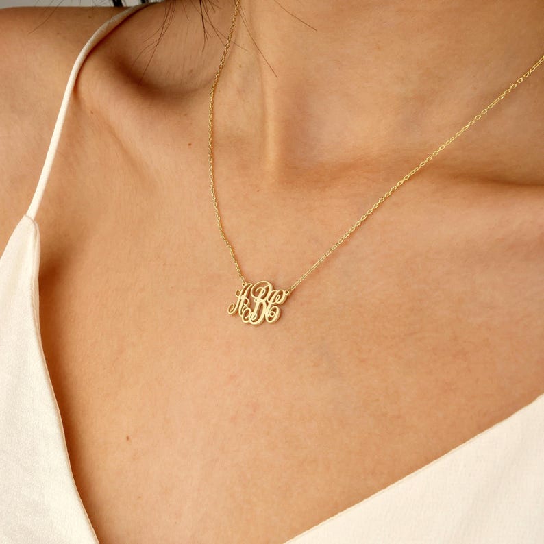 May include: A gold-colored monogram necklace with the letters "ABC" in a delicate, cursive font. The necklace is displayed on a person's chest, showcasing the pendant's design and the chain's subtle elegance. The necklace is a personalized jewelry item.