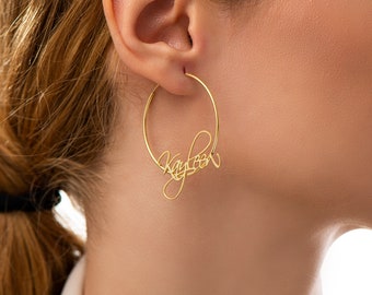 May include: Gold-colored hoop earrings with the name "Kayleen" written in a cursive script. The earrings are circular and appear to be made of a shiny metal. The name is integrated into the hoop design.