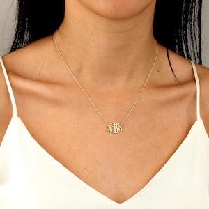 May include: A gold-colored monogram necklace with a delicate chain. The pendant features three stylized initials. The necklace is worn against a white top.