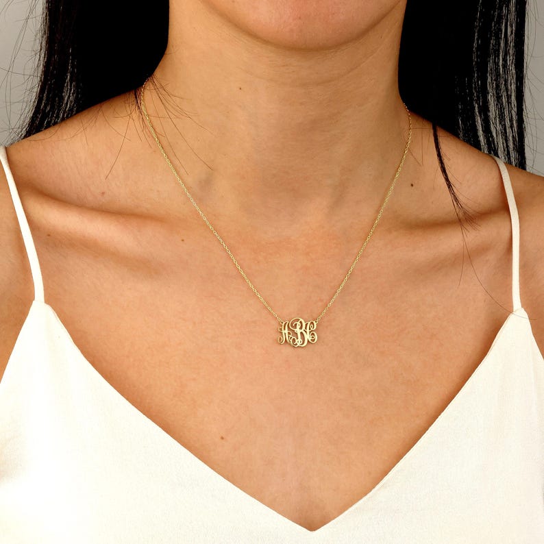 May include: A gold-colored monogram necklace with a delicate chain. The pendant features three stylized initials. The necklace is worn against a white top.