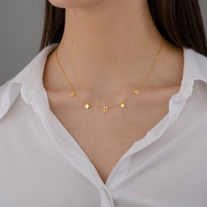 May include: A gold necklace with a delicate chain and five charms: four letters and one star. The letters are "S", "D", "B", and a fourth letter that is unclear. The necklace is worn by a person with a white shirt.