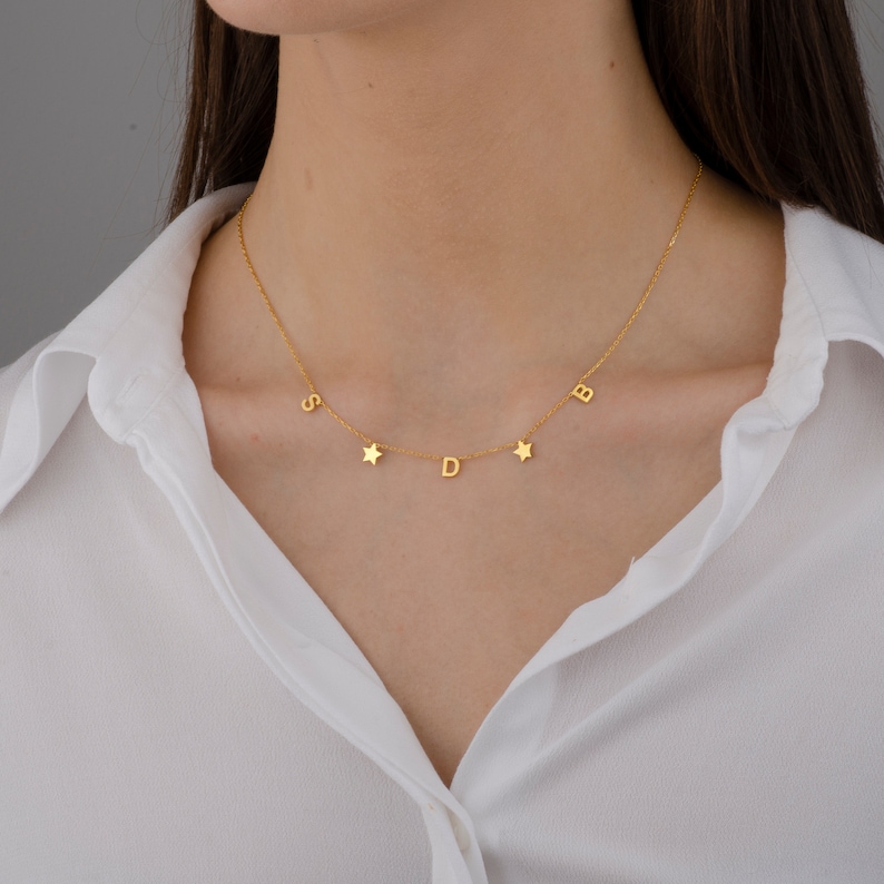 May include: A gold necklace with a delicate chain and five charms: four letters and one star. The letters are "S", "D", "B", and a fourth letter that is unclear. The necklace is worn by a person with a white shirt.