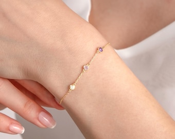 May include: A delicate gold chain bracelet featuring three small, bezel-set circular gemstones spaced evenly along the length. The stones include a clear white gem, a light pink gem, and a vibrant purple gem, all set in polished gold metal.