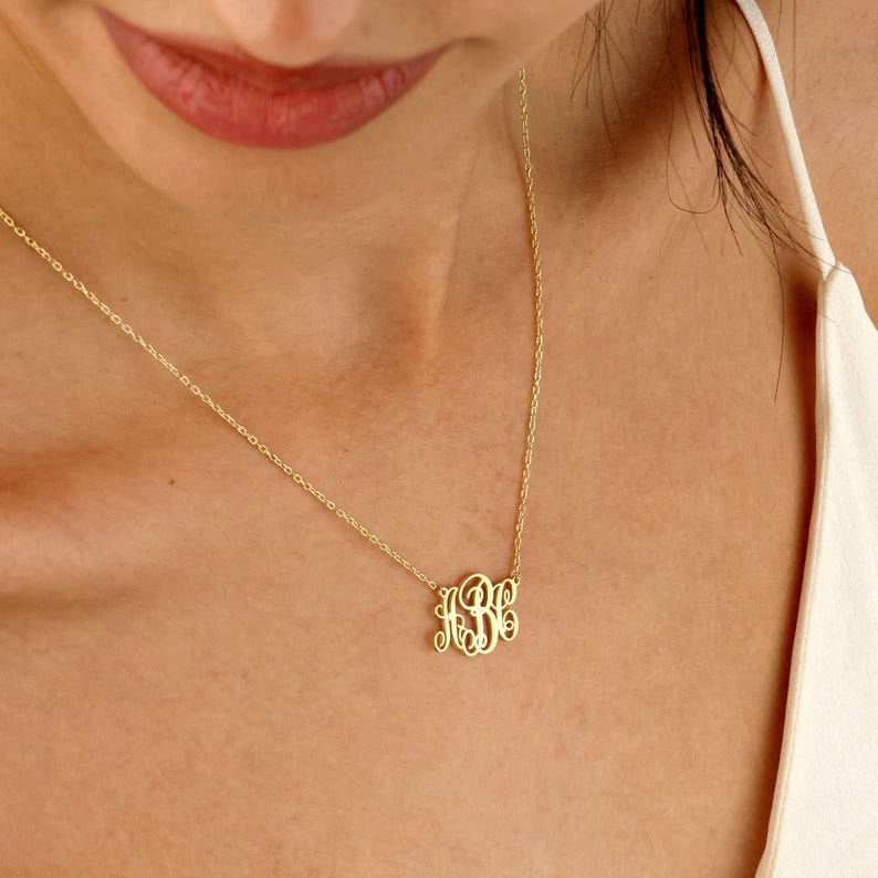 May include: A gold-colored monogram necklace with the initials "KBE" in a delicate, cursive font. The necklace hangs on a fine chain, resting on a person's chest. The background is a soft, neutral tone, highlighting the jewelry.
