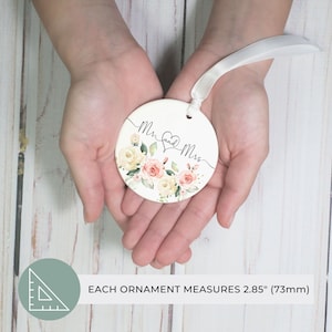K&ouml;nnte beinhalten: Runder, wei&szlig;er Ornament mit den Worten "Mr. and Mrs." in eleganter Schrift, verziert mit einem Herz und floralen Designs in zarten Rosa-, Pfirsich- und Cremet&ouml;nen. Das Ornament hat ein wei&szlig;es Band zum Aufh&auml;ngen. Jedes Ornament misst 73 mm.