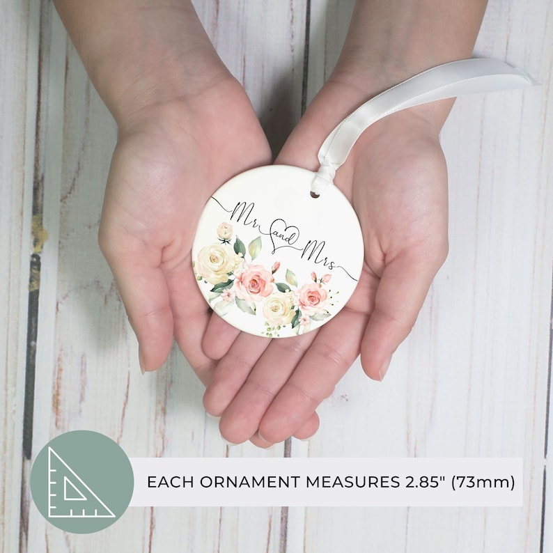 K&ouml;nnte beinhalten: Runder, wei&szlig;er Ornament mit den Worten "Mr. and Mrs." in eleganter Schrift, verziert mit einem Herz und floralen Designs in zarten Rosa-, Pfirsich- und Cremet&ouml;nen. Das Ornament hat ein wei&szlig;es Band zum Aufh&auml;ngen. Jedes Ornament misst 73 mm.