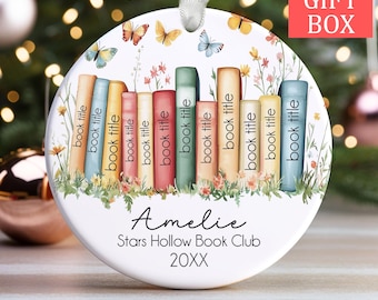 Può includere: Un ornamento natalizio rotondo e bianco con un design di libreria colorata, farfalle e fiori. L'ornamento presenta il testo "Amelie", "Stars Hollow Book Club" e "20XX". Un nastro bianco è attaccato per appenderlo. Le parole "FREE GIFT BOX" sono nell'angolo in alto a destra.