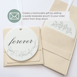 K&ouml;nnte beinhalten: Wei&szlig;es Keramik-Scheibenornament mit dem Wort "forever" in schwarzer Schrift, umgeben von einem floralen Design. Es h&auml;ngt an einem wei&szlig;en Band und befindet sich in einem beigefarbenen Wildlederbeutel. Ein weiteres Ornament mit floralem Design ist ebenfalls sichtbar.