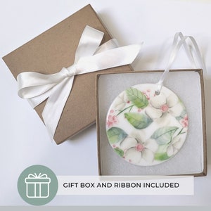 K&ouml;nnte beinhalten: Runder Keramikschmuck mit floralem Design aus wei&szlig;en Magnolienbl&uuml;ten, gr&uuml;nen Bl&auml;ttern und rosa Akzenten. Pr&auml;sentiert in einer braunen Geschenkbox mit wei&szlig;em Band. Der Text "GIFT BOX AND RIBBON INCLUDED" ist ebenfalls sichtbar.