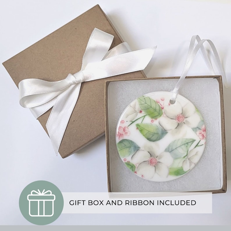 K&ouml;nnte beinhalten: Runder Keramikschmuck mit floralem Design aus wei&szlig;en Magnolienbl&uuml;ten, gr&uuml;nen Bl&auml;ttern und rosa Akzenten. Pr&auml;sentiert in einer braunen Geschenkbox mit wei&szlig;em Band. Der Text "GIFT BOX AND RIBBON INCLUDED" ist ebenfalls sichtbar.