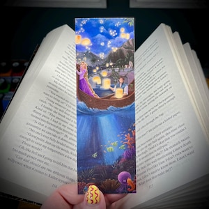 Peut inclure: Marque-page rectangulaire illustrant une scène de conte de fées. On y voit un couple dans une barque, entouré de lanternes flottantes sous un ciel étoilé. Le marque-page est inséré entre les pages d'un livre.
