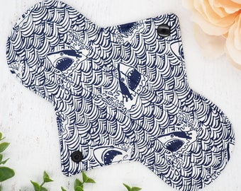 May include: A white reusable cloth pad with a blue and white shark print. The pad has two black snaps on the sides.
