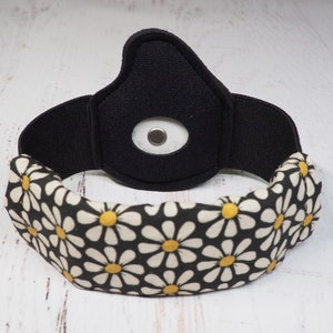 Op de afbeelding: Zwart hoofdband met een witte en gele madeliefjesprint. Het hoofdband heeft een zwarte stoffen patch met een kleine ronde opening in het midden.