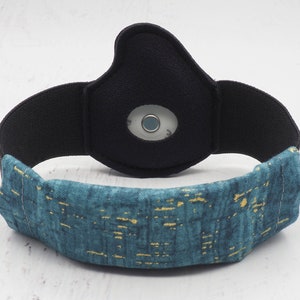 Op de afbeelding: Een teal en gouden gepatroneerd hoofdband met een zwart elastisch band en een ronde, zwarte sensor in het midden.