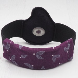 Op de afbeelding: Een paarse stoffen hoofdband met een zwart elastisch band en een klein rond sensorvenster. De stof heeft een patroon van kleine grijze bladeren.