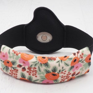 May include: A black and white floral fabric cover for a continuous glucose monitor (CGM). The cover has a white background with pink and orange flowers and green leaves. The CGM is visible through a hole in the black fabric.