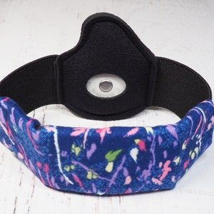 Op de afbeelding: Een zwart en blauw hoofdband met een bloemenpatroon. Het hoofdband heeft een ingebouwde zak voor een klein apparaat, zoals een fitness tracker.