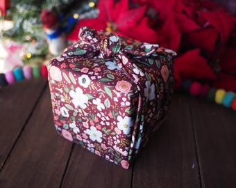 Puede incluir: Un regalo cuadrado envuelto en una tela floral con flores blancas, rosas y amarillas sobre un fondo burdeos. La tela está atada en la parte superior con un lazo a juego. El regalo está sobre una superficie de madera oscura.
