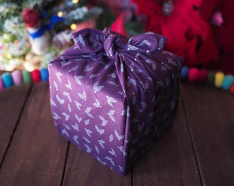 Puede incluir: Un regalo cuadrado envuelto en una tela morada oscura con un estampado de hojas blancas. La tela está atada en la parte superior, creando un nudo decorativo. El regalo está colocado sobre una superficie de madera, con una guirnalda colorida en el fondo.