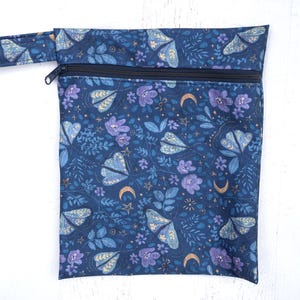 May include: A rectangular fabric pouch with a black zipper closure and a matching carry strap. The pouch features a dark blue background printed with a pattern of light blue moths, purple flowers, gold crescent moons, and small gold stars.