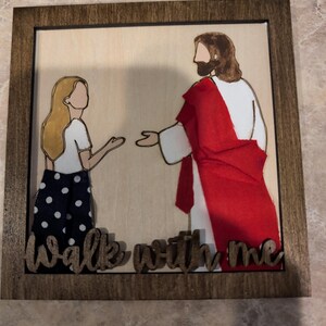 Pode incluir: Uma placa de parede quadrada de madeira com fundo de madeira clara e uma ilustra&ccedil;&atilde;o em camadas de uma pessoa com cabelo loiro e uma figura com um manto vermelho. O texto "Walk with me" est&aacute; recortado em madeira escura na base.