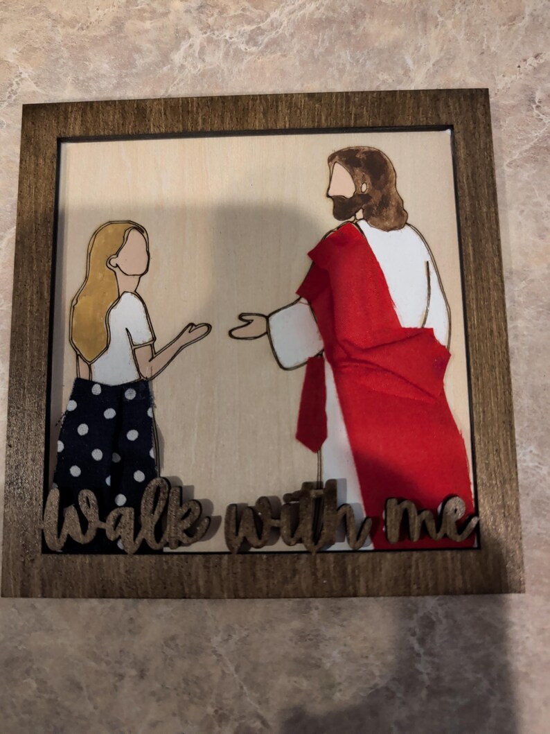 Pode incluir: Uma placa de parede quadrada de madeira com fundo de madeira clara e uma ilustra&ccedil;&atilde;o em camadas de uma pessoa com cabelo loiro e uma figura com um manto vermelho. O texto "Walk with me" est&aacute; recortado em madeira escura na base.