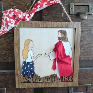 Pode incluir: Um enfeite de parede quadrado de madeira com uma figura de cabelos loiros longos, camisa branca e saia azul de bolinhas, de frente para uma figura de t&uacute;nica branca com faixa de tecido vermelho. O texto "Walk with me" est&aacute; na base, com la&ccedil;o.