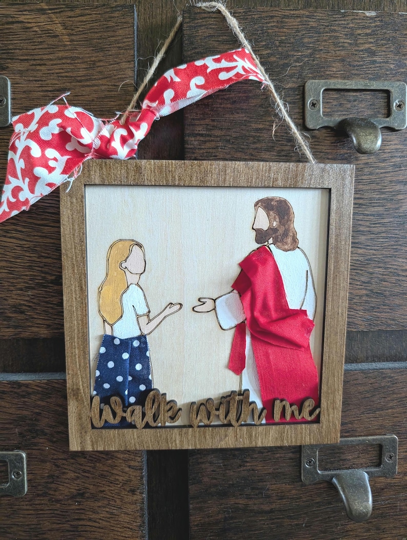 Pode incluir: Um enfeite de parede quadrado de madeira com uma figura de cabelos loiros longos, camisa branca e saia azul de bolinhas, de frente para uma figura de t&uacute;nica branca com faixa de tecido vermelho. O texto "Walk with me" est&aacute; na base, com la&ccedil;o.