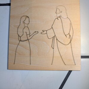 Pode incluir: Uma placa de madeira clara de formato quadrado com um desenho de linha preta de uma pessoa com cabelo comprido em frente a uma figura de t&uacute;nica com cabelo pelos ombros. As duas figuras est&atilde;o viradas uma para a outra com as m&atilde;os estendidas.
