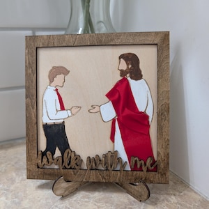 Pode incluir: Uma placa de madeira quadrada com uma ilustra&ccedil;&atilde;o cortada a laser de uma pessoa de camisa branca e cal&ccedil;as pretas diante de uma figura com t&uacute;nica branca e faixa vermelha. O texto "walk with me" est&aacute; recortado na madeira na parte inferior.