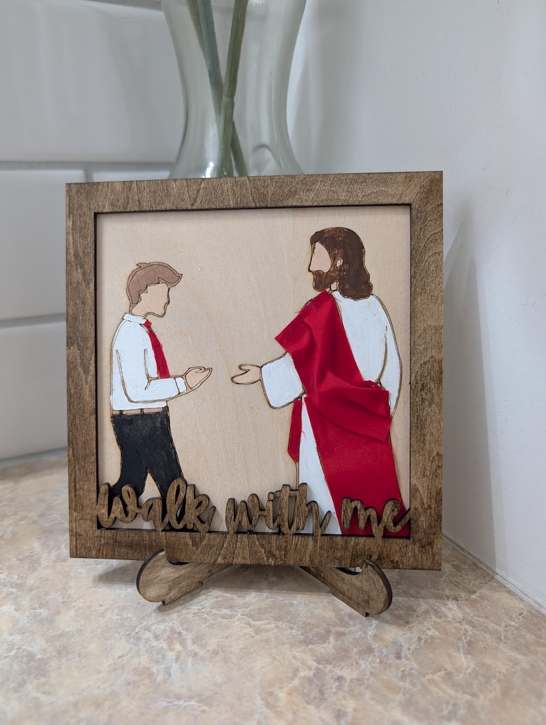 Pode incluir: Uma placa de madeira quadrada com uma ilustra&ccedil;&atilde;o cortada a laser de uma pessoa de camisa branca e cal&ccedil;as pretas diante de uma figura com t&uacute;nica branca e faixa vermelha. O texto "walk with me" est&aacute; recortado na madeira na parte inferior.