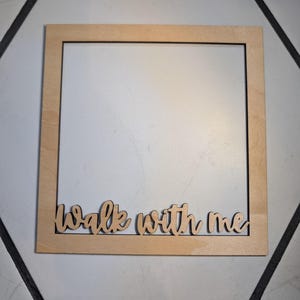 Pode incluir: Uma moldura quadrada de madeira de cor clara com acabamento liso. A borda inferior da moldura apresenta a frase "walk with me" recortada em uma fonte cursiva, integrada diretamente na borda de madeira da estrutura quadrada.