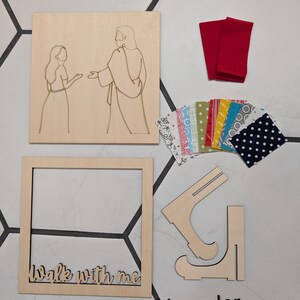 Pode incluir: Um kit de artesanato em madeira com um painel quadrado com uma ilustra&ccedil;&atilde;o de linhas de duas figuras, uma moldura quadrada com o texto "Walk with me", duas pe&ccedil;as de suporte em madeira, dois peda&ccedil;os de tecido vermelho e retalhos coloridos.