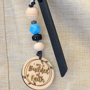 May include: A wooden keychain with the phrase "I am a Builder of Faith" engraved on a round wooden charm. The keychain features a series of beads in beige, blue, black, and white, and is attached to a black leather strap.