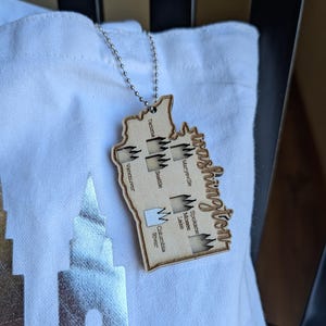 May include: A light-colored wooden ornament shaped like the state of Washington, featuring a silver ball chain. The surface is laser-engraved with the word Washington, city names including Tacoma, Seattle, and Spokane, and several mountain icons.