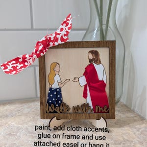 Pode incluir: Uma moldura de madeira quadrada contendo um painel claro com a representa&ccedil;&atilde;o de uma pessoa e uma figura com t&uacute;nica vermelha. O texto "walk with me" est&aacute; na parte inferior. Um la&ccedil;o de tecido com padr&atilde;o vermelho e branco est&aacute; preso ao topo.