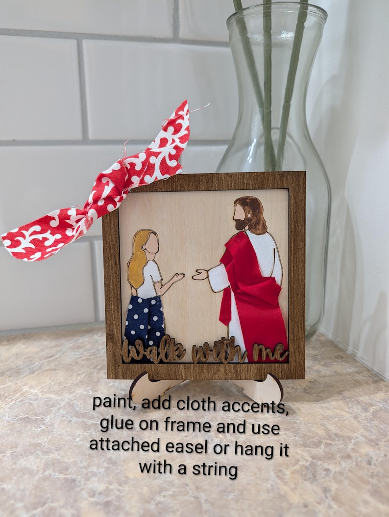 Pode incluir: Uma moldura de madeira quadrada contendo um painel claro com a representa&ccedil;&atilde;o de uma pessoa e uma figura com t&uacute;nica vermelha. O texto "walk with me" est&aacute; na parte inferior. Um la&ccedil;o de tecido com padr&atilde;o vermelho e branco est&aacute; preso ao topo.