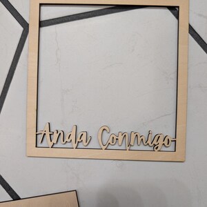 Pode incluir: Uma moldura quadrada de madeira clara com o texto "Anda Conmigo" cortado a laser fixado na borda inferior. A moldura tem um acabamento de madeira natural lisa e um design simples com um centro oco para exibir objetos.
