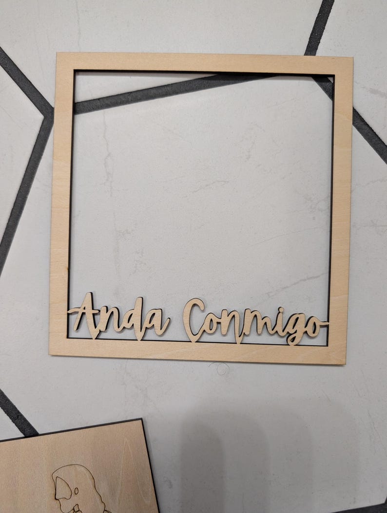 Pode incluir: Uma moldura quadrada de madeira clara com o texto "Anda Conmigo" cortado a laser fixado na borda inferior. A moldura tem um acabamento de madeira natural lisa e um design simples com um centro oco para exibir objetos.