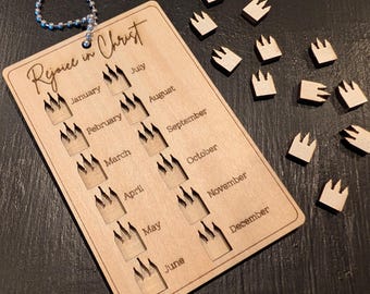 May include: A rectangular wooden calendar tag with a metal ball chain, engraved with the text "Rejoice in Christ" and twelve crown-shaped cutouts labeled with months from January to December, accompanied by several small wooden crown tokens.