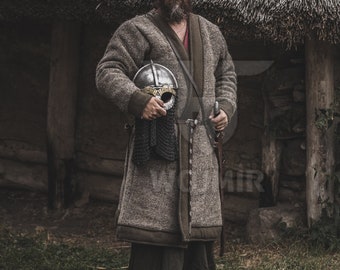 May include: A man wearing a brown fur coat and holding a metal helmet stands in front of a thatched roof hut. He is wearing a red shirt underneath the coat and has a beard.